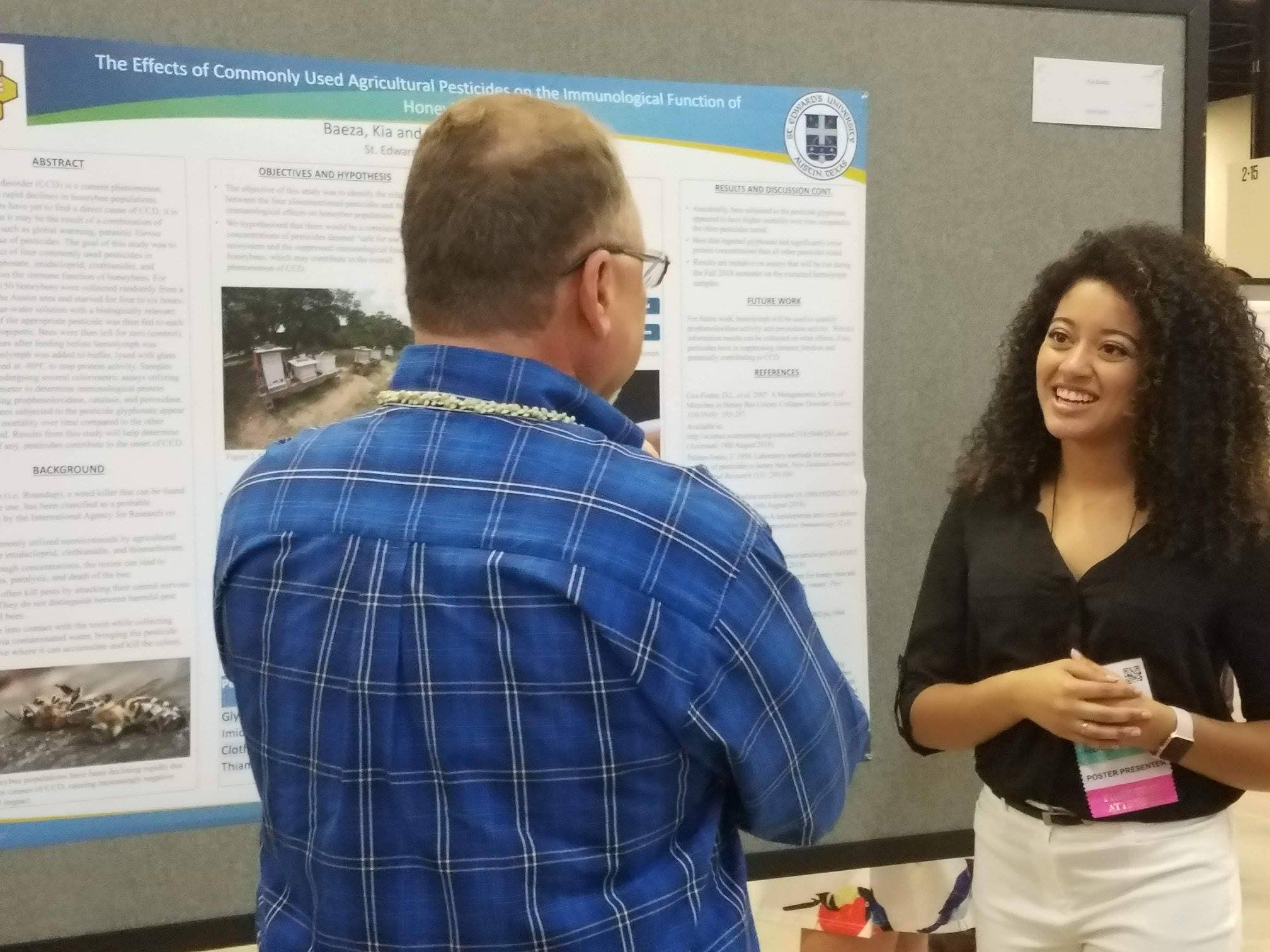A smiling woman presents a scientific poster about pesticides and honeybees to a man.